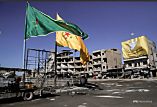 Place Al-Naim à Al-Raqqah. Droite : Drapeau géant des FDS accroché sur la façade d'un bâtiment. Gauche : Drapeaux géant des organisations kurdes en Syrie (GPJ et YPJ), principales composantes des FDS (Compte Twitter, 18 octobre 2017)