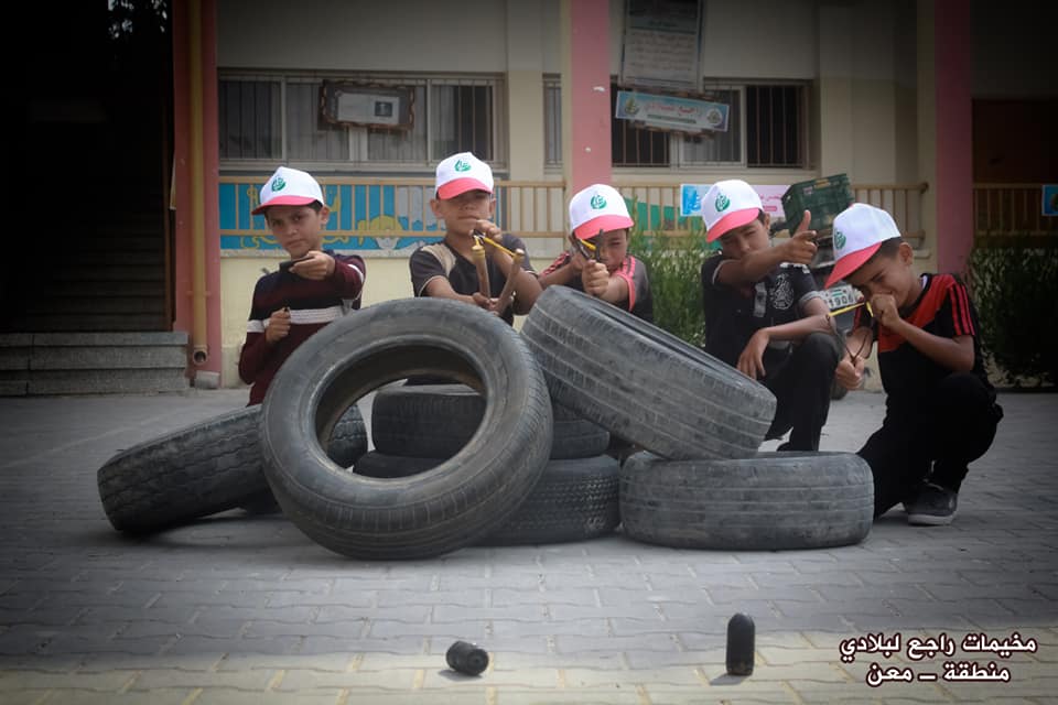 Using slingshots while hiding behind tires in the summer camp in the Ma'an region – Abu Obeida branch in Khan Yunis (Facebook page of the committee of the summer camps in the Ma'an region, July 15, 2018).