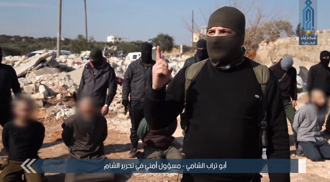 Abu Turab al-Suri, senior security operative of the Headquarters for the Liberation of Al-Sham. ISIS operatives are seen in the background, before being executed. Behind them are the ruins of the command post of the Headquarters for the Liberation of Al-Sham (Ibaa, January 20, 2019).