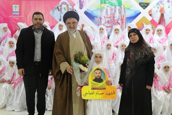 Group ceremony of girls coming of age at the Al-Mahdi schools in Mashghara, Kafr Fila and Al-Ahmadiyah. A total of 88 girls coming of age from these schools took part in the ceremony (Facebook page of the Al-Mahdi school in Mashghara, February 20-24, 2019). The girl in the middle is holding a photo of a shahid.