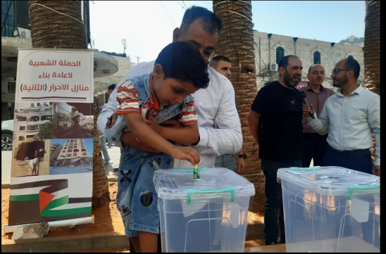 A child puts his donation into the box. Mazen al-Danbak stands in back to the right, giving an interview (aljazeera.net, July 30, 2023).